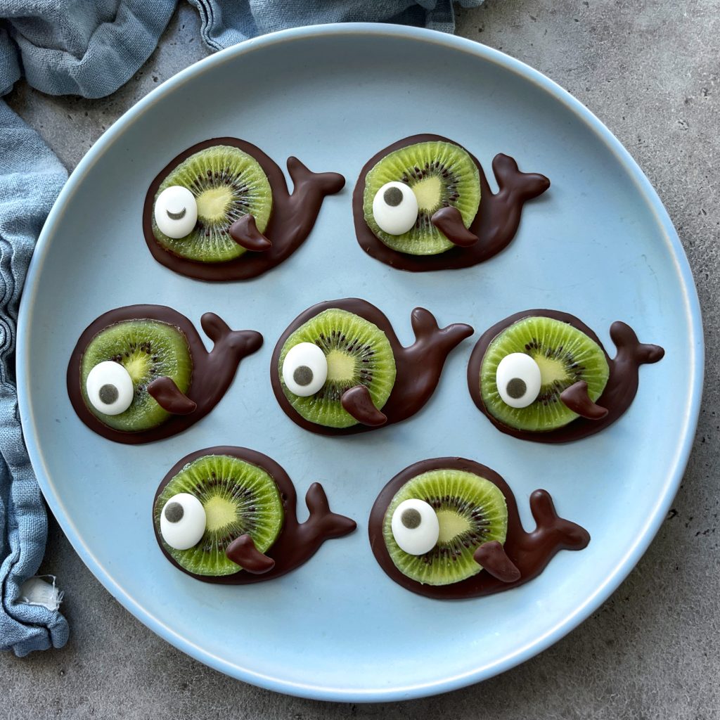 A plate with seven fish-shaped bites made from chocolate, slices of kiwi, and candy eyes, arranged neatly on a light blue plate—perfect for fun food recipes or creative dishes.