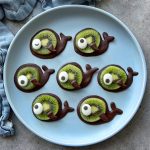 A plate with seven fish-shaped bites made from chocolate, slices of kiwi, and candy eyes, arranged neatly on a light blue plate—perfect for fun food recipes or creative dishes.