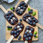 Five chocolate-dipped popsicle bites on a wooden board, topped with fresh blueberries and candy eyes, garnished with basil leaves—these playful food recipes create a fun, whimsical look perfect for health-conscious treats.