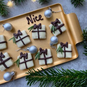 A gold tray holds white chocolate bites decorated as gift boxes with chocolate bows and sprigs of rosemary. Surrounded by silver Christmas ornaments and green pine branches, this festive food dish spells out "Nice" in chocolate.