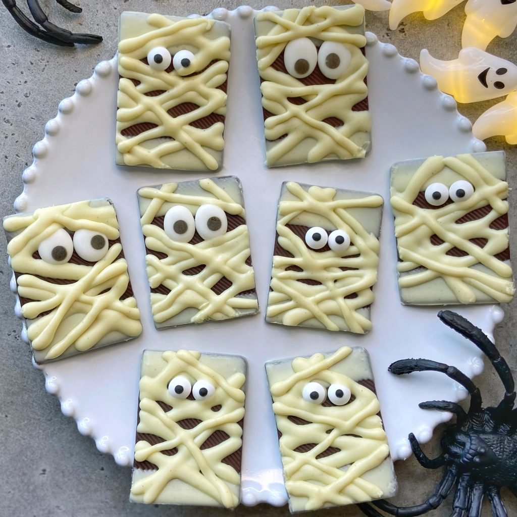 A plate of flat, rectangular chocolate pieces decorated as mummies with white chocolate bandages and candy eyes. Plastic spider and ghost decorations surround the plate.