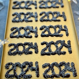 A gold tray displays several chocolate decorations shaped as “2024,” each topped with small silver sprinkles, perfect for adding a festive touch to your party bites or inspiring creative dessert recipes.