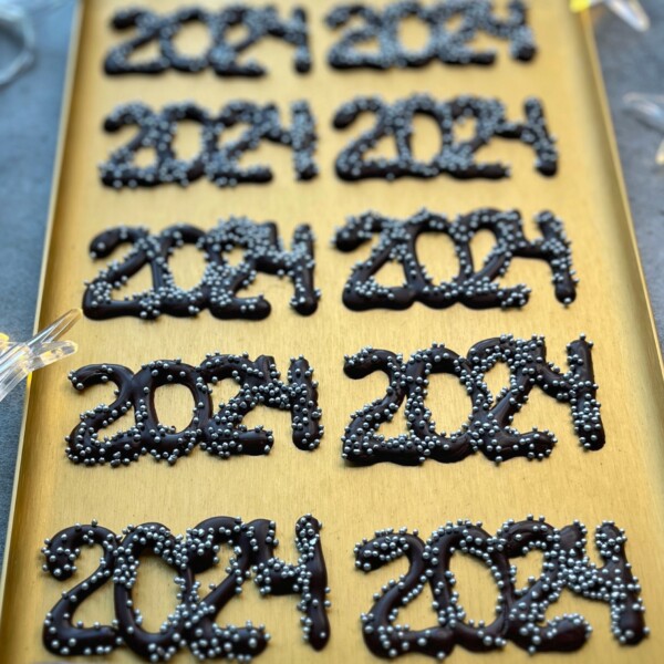 A gold tray displays several chocolate decorations shaped as “2024,” each topped with small silver sprinkles, perfect for adding a festive touch to your party bites or inspiring creative dessert recipes.