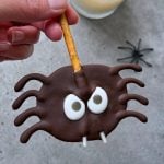 A hand holds a chocolate-covered spider-shaped treat with pretzel legs, candy eyes, and two white fangs—a fun food recipe—while a plastic spider and a glass of milk rest in the background on a gray surface.