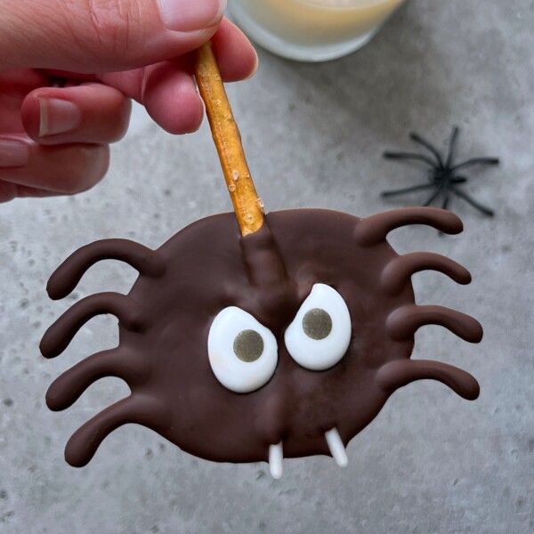A hand holds a chocolate-covered spider-shaped treat with pretzel legs, candy eyes, and two white fangs—a fun food recipe—while a plastic spider and a glass of milk rest in the background on a gray surface.