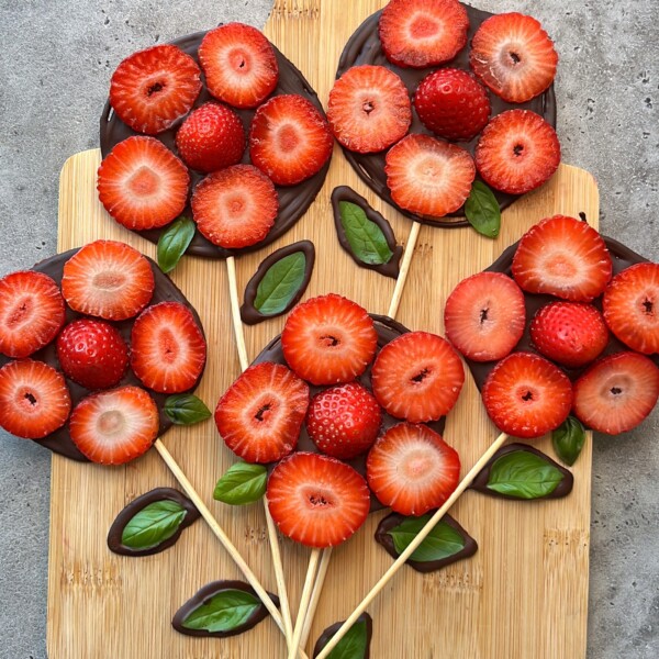Five lollipop-shaped bites made of chocolate disks topped with sliced strawberries and basil leaves, arranged on wooden skewers to resemble flower bouquets, are displayed on a wooden board—perfect for adding creativity to your food recipes.