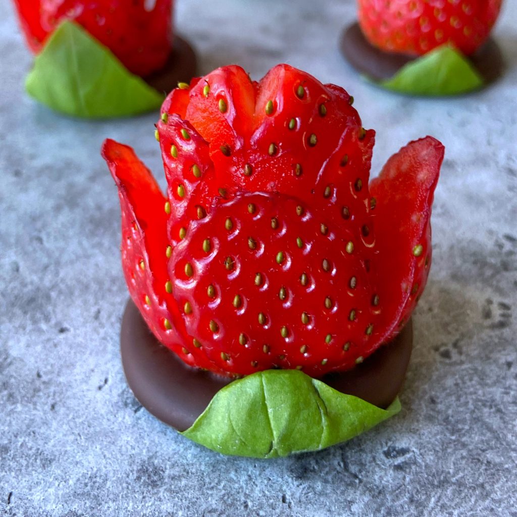 A strawberry carved to resemble a flower sits on a round chocolate base with a green leaf, evoking the look of chocolate strawberry roses. Other similar strawberry flowers are visible in the background on a gray surface.