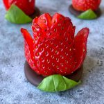 A chocolate strawberry rose, carved to resemble a blooming flower, sits atop a round piece of chocolate with a green leaf at the base, displayed on a gray surface. Other chocolate strawberry roses are softly blurred in the background.