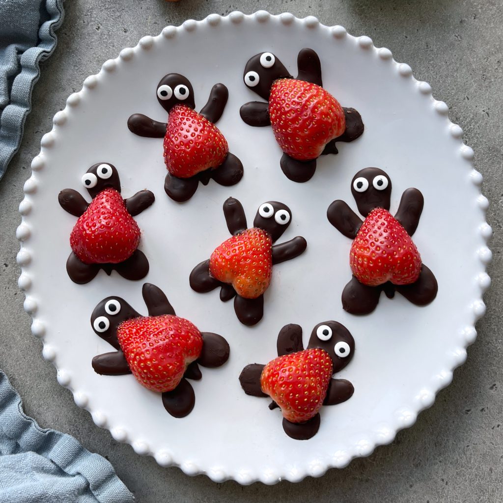 A white plate with seven adorable chocolate turtles with strawberry shells, featuring strawberries as shells and chocolate forming the heads, legs, and tails—complete with edible eyes for a delightful treat.