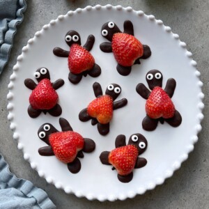 A white plate with seven adorable chocolate turtles with strawberry shells, featuring strawberries as shells and chocolate forming the heads, legs, and tails—complete with edible eyes for a delightful treat.