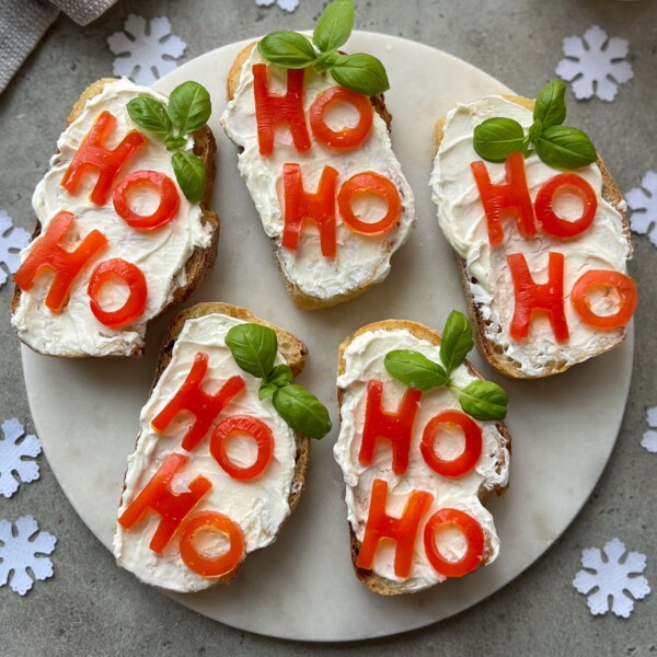 Five slices of Christmas Bruschetta with cream cheese are topped with red bell pepper pieces spelling “HO HO” and garnished with basil leaves, arranged on a round plate surrounded by festive paper snowflakes.