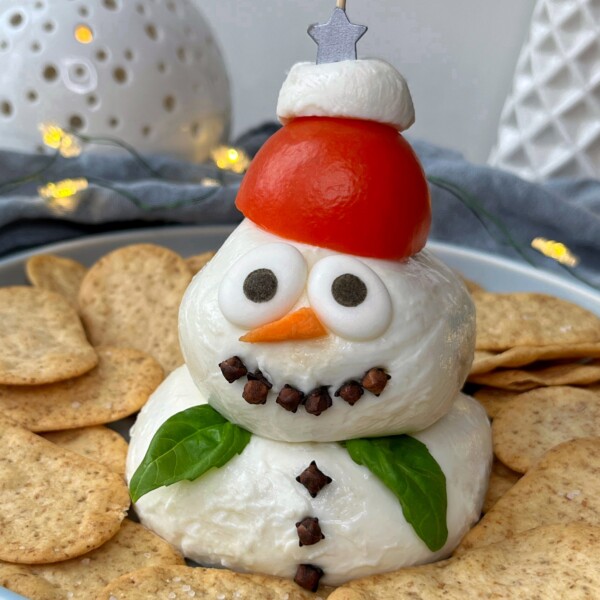 A snowman-shaped cheese ball appetizer, perfect for festive bites, is decorated with black olive eyes, a carrot nose, clove buttons and mouth, basil leaf arms, a tomato-and-mozzarella hat, and surrounded by round crackers.
