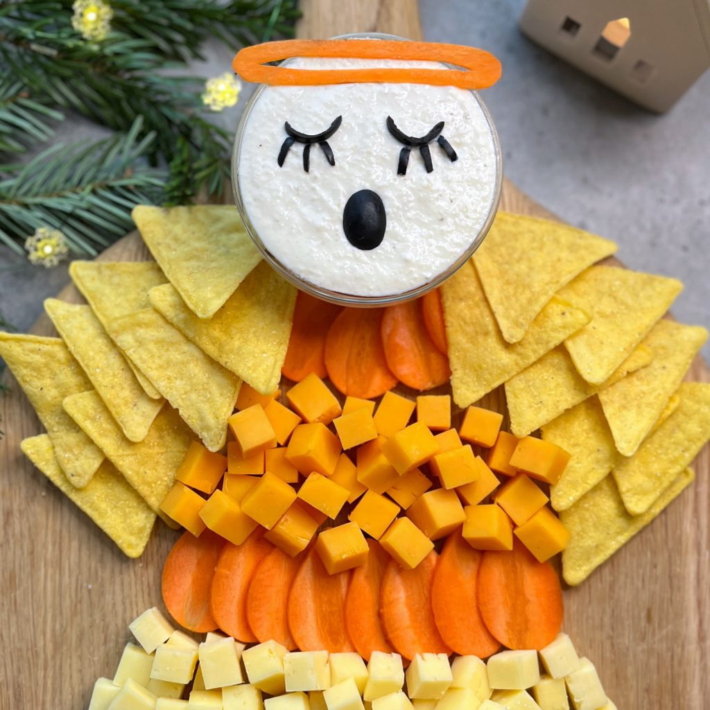 A festive snack platter arranged to look like an angel, featuring healthy bites like cheese cubes and carrot slices, a dip for the head, and tortilla chips for wings. Decor and pine branches appear in the background.