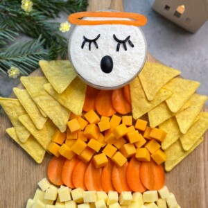 A festive snack platter arranged to look like an angel, featuring healthy bites like cheese cubes and carrot slices, a dip for the head, and tortilla chips for wings. Decor and pine branches appear in the background.