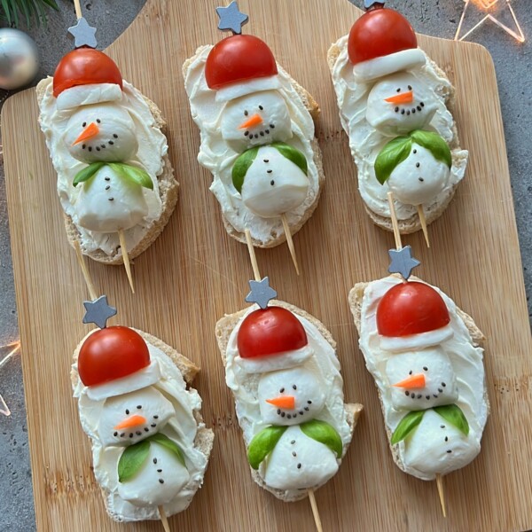 Six slices of bread topped with cream cheese, shaped like snowmen using cherry tomatoes for hats and veggies for details—these festive bites make delightful Christmas Party Appetizers, beautifully arranged on a wooden board.