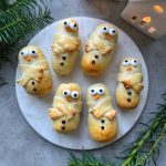 Six cute Christmas Sausage Rolls decorated as snowmen with candy eyes, orange candy noses, and chocolate buttons, arranged on a round white plate with greenery and a lit white house decoration nearby.