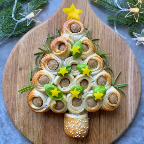 A festive Christmas Tree Sausage Rolls arrangement, shaped like a tree with sausage rolls as ornaments, topped with yellow star-shaped veggies and green leaves, displayed on a wooden board surrounded by holiday lights and pine branches.