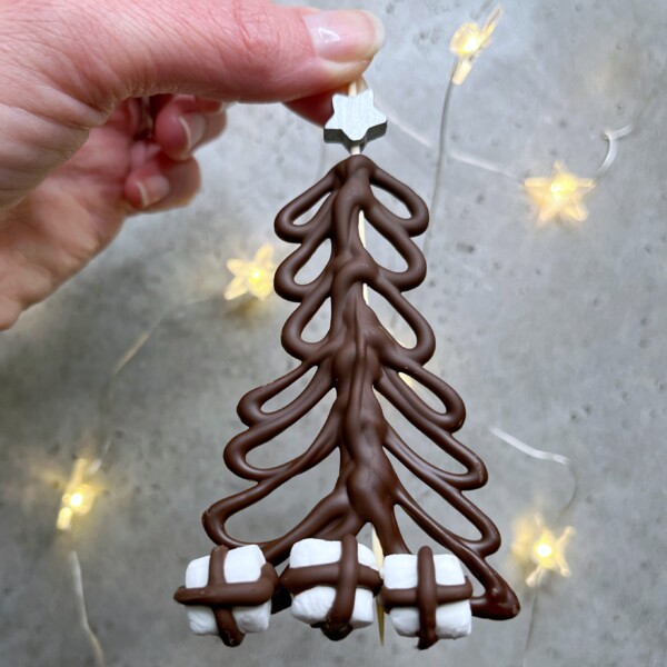 A hand holds a Christmas tree-shaped chocolate decoration with a silver star on top and marshmallows at the base—perfect for festive bites or adding fun to your holiday food recipes. Star-shaped fairy lights are blurred in the background.