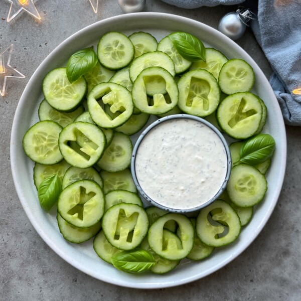 A festive Christmas Veggie Tray features cucumber slices arranged in a wreath, with some spelling MERRY XMAS. Fresh basil leaves and a bowl of white dipping sauce sit at the center, surrounded by decorative lights and ornaments.