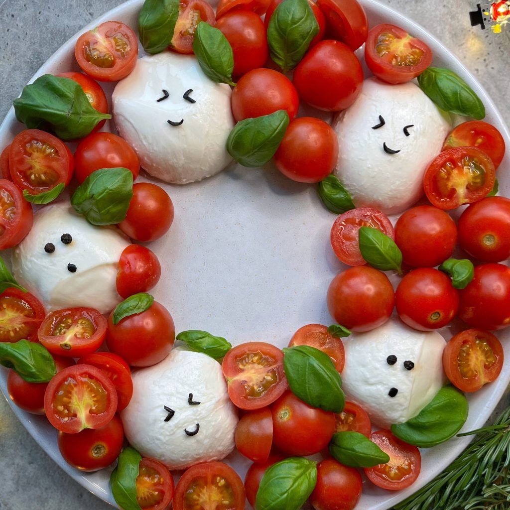 A festive plate arranged like a holiday wreath with mozzarella balls decorated as smiling faces, surrounded by cherry tomatoes and fresh basil leaves.