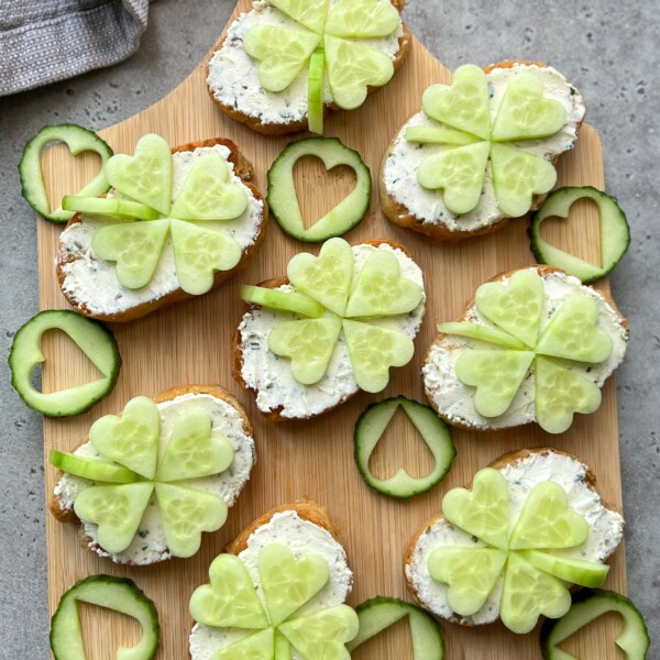 Small slices of bread with herb cream cheese are topped with cucumber pieces arranged as shamrocks. These festive bites make a charming addition to your snack display, blending fun with health for creative party food ideas.