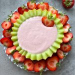 A round honeydew melon rind filled with creamy pink cream cheese fruit dip sits on a white plate, surrounded by halved fresh strawberries. One whole strawberry is placed on the edge of the melon rind.