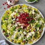 A plate of Crispy Prosciutto Salad topped with pomegranate seeds, chopped cucumber, diced apple, fresh mint leaves, and creamy dressing. Garlic, mint, and a striped napkin surround the plate on a gray surface.