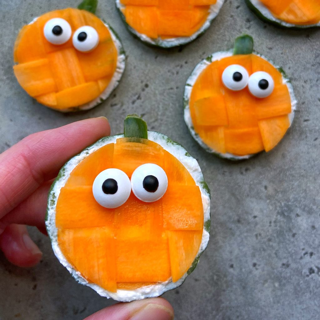 Round cucumber bites decorated to look like pumpkins with carrot pieces, candy eyes, and green cucumber skin tops, arranged on a gray surface.
