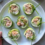 A plate of cucumber roll-ups filled with a creamy salmon and avocado mixture, garnished with green herbs, makes for delicious health bites neatly arranged on a light-colored ceramic dish.