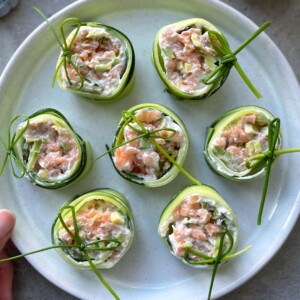A plate of cucumber roll-ups filled with a creamy salmon and avocado mixture, garnished with green herbs, makes for delicious health bites neatly arranged on a light-colored ceramic dish.
