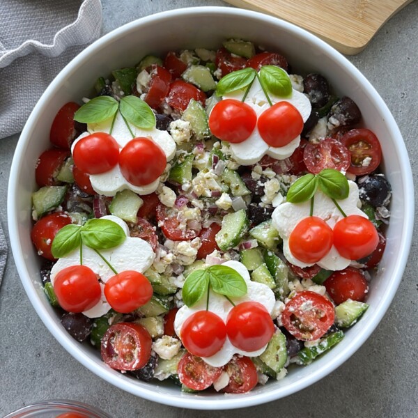 A bowl of cucumber tomato feta salad topped with clusters of mozzarella slices, cherry tomatoes, and fresh basil leaves arranged to look like cherries. The salad contains tomatoes, cucumbers, onions, olives, and feta.