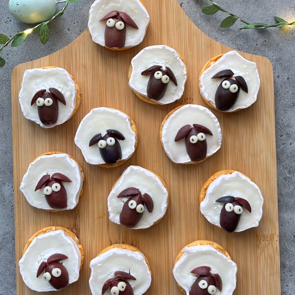 Whimsical round cracker bites topped with a white spread, decorated as playful sheep faces using black olives and cheese for eyes—a fun addition to your food recipes or party dishes.