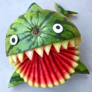 A watermelon carved to look like a shark, with a gaping mouth of red fruit bites, white teeth, and eyes made from round pieces of fruit and chocolate, plus a tail fin cut from the rind—a fun addition to summer food recipes.