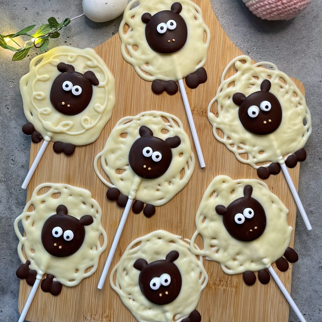 Seven sheep-shaped white and dark chocolate lollipops with cute faces and big eyes are arranged on a wooden board, resembling cartoon sheep with textured wool and ears—adorable bites perfect for fun food recipes.