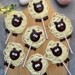 Seven sheep-shaped white and dark chocolate lollipops with cute faces and big eyes are arranged on a wooden board, resembling cartoon sheep with textured wool and ears—adorable bites perfect for fun food recipes.