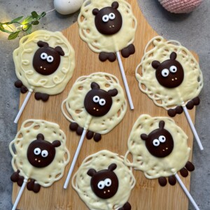 Seven sheep-shaped white and dark chocolate lollipops with cute faces and big eyes are arranged on a wooden board, resembling cartoon sheep with textured wool and ears—adorable bites perfect for fun food recipes.
