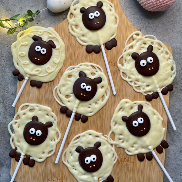 Seven sheep-shaped white and dark chocolate lollipops with cute faces and big eyes are arranged on a wooden board, resembling cartoon sheep with textured wool and ears—adorable bites perfect for fun food recipes.