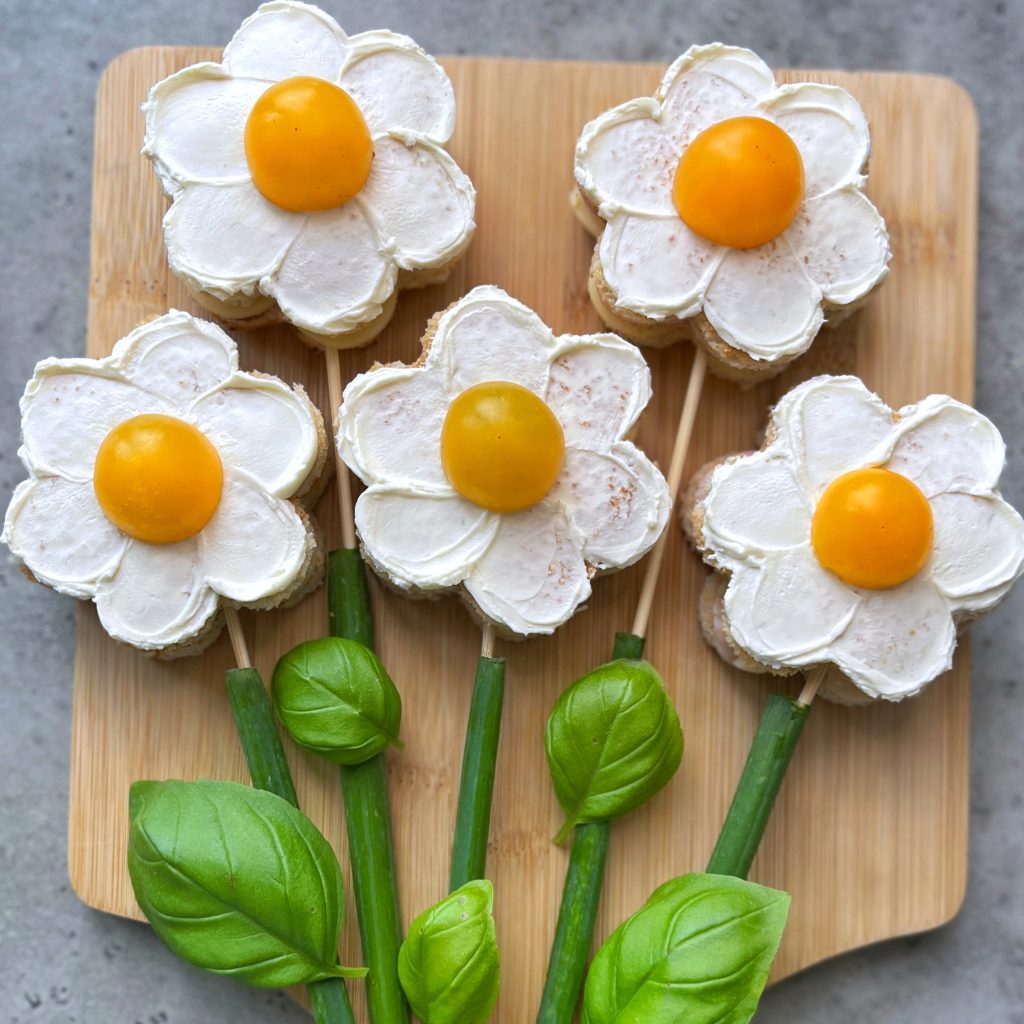 Five flower-shaped bites with white bread and cream petals, yellow cherry tomato centers, and green onion stems with basil leaves, arranged on a wooden board to resemble flowers—perfect for creative food recipes or healthy snacks.