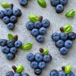 Clusters of blueberries arranged in the shape of grape bunches, with small green leaves and chocolate accents on a speckled gray surface—perfect for health-focused bites or creative food recipes.