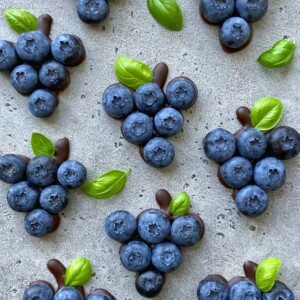 Clusters of blueberries arranged in the shape of grape bunches, with small green leaves and chocolate accents on a speckled gray surface—perfect for health-focused bites or creative food recipes.