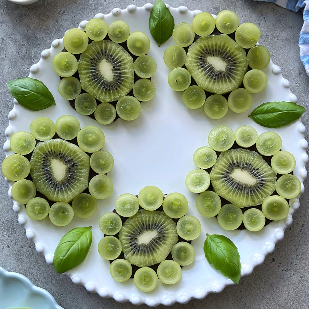 A white plate displays four dark chocolate kiwi flowers, with sliced kiwi centers and halved green grapes as petals, garnished with fresh basil leaves.