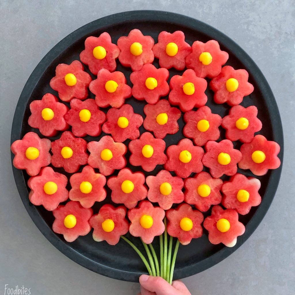 A round black tray holds watermelon pieces cut into flower shapes with yellow mango centers, arranged like a bouquet with green stems made from cut basil.