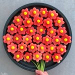 A round tray displays watermelon slices cut into flower shapes with yellow centers, arranged like a bouquet with green stems—perfect for healthy bites and food recipes—with a hand holding the stems at the bottom.