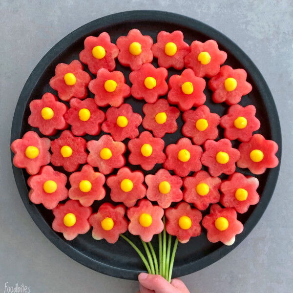 A round tray displays watermelon slices cut into flower shapes with yellow centers, arranged like a bouquet with green stems—perfect for healthy bites and food recipes—with a hand holding the stems at the bottom.