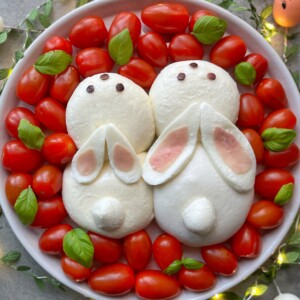 A playful dish featuring two mozzarella balls shaped like bunnies, with tomato ears and chocolate chip eyes, surrounded by cherry tomatoes and basil leaves—an adorable idea for fun bites in creative food recipes.