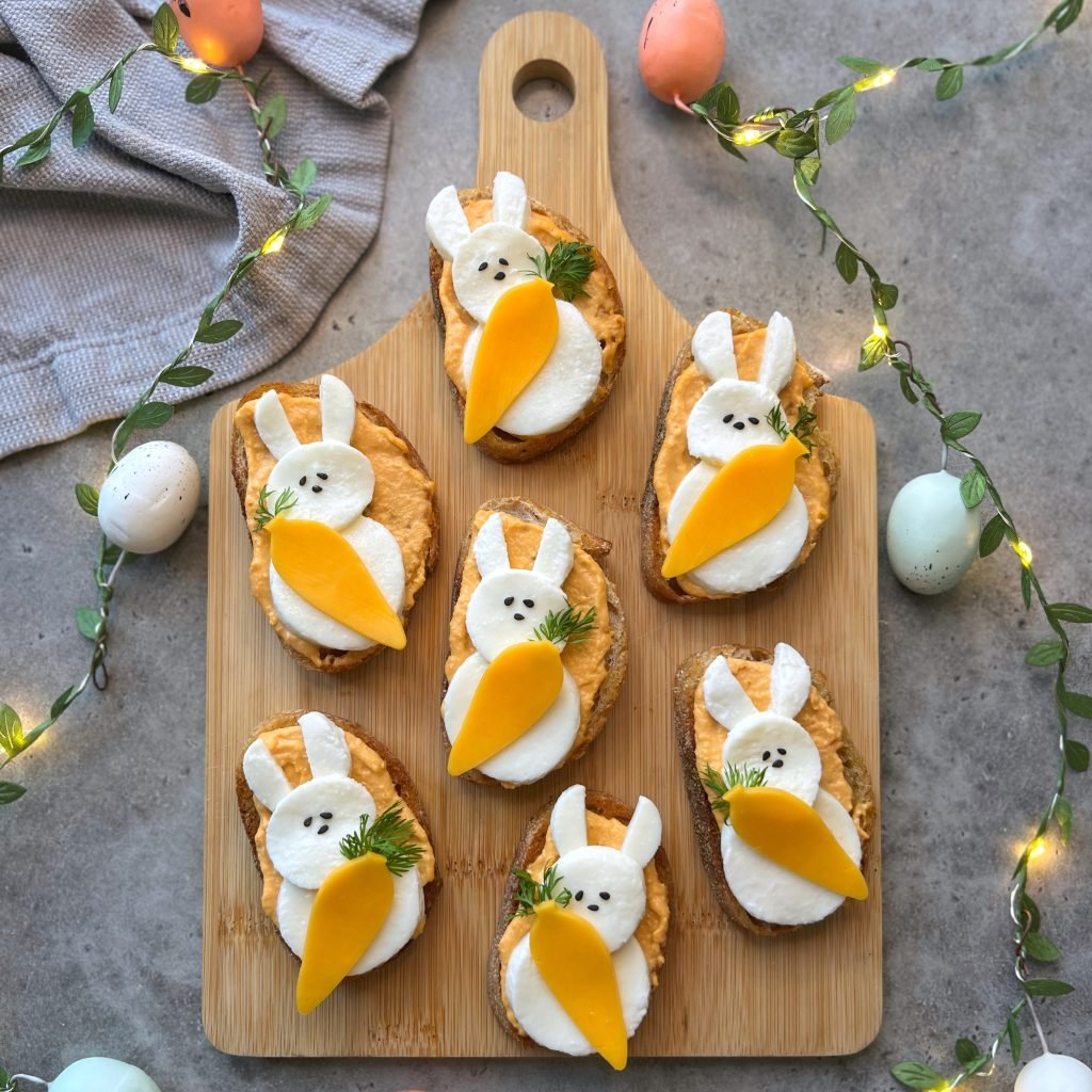 A wooden board holds eight bunny-shaped bites with white cheese, carrot slices, and black sesame eyes, surrounded by string lights, pastel eggs, and a gray cloth—perfect for adding fun to your food recipes.