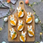 A wooden board holds eight bunny-shaped bites with white cheese, carrot slices, and black sesame eyes, surrounded by string lights, pastel eggs, and a gray cloth—perfect for adding fun to your food recipes.