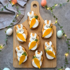 A wooden board holds eight bunny-shaped bites with white cheese, carrot slices, and black sesame eyes, surrounded by string lights, pastel eggs, and a gray cloth—perfect for adding fun to your food recipes.
