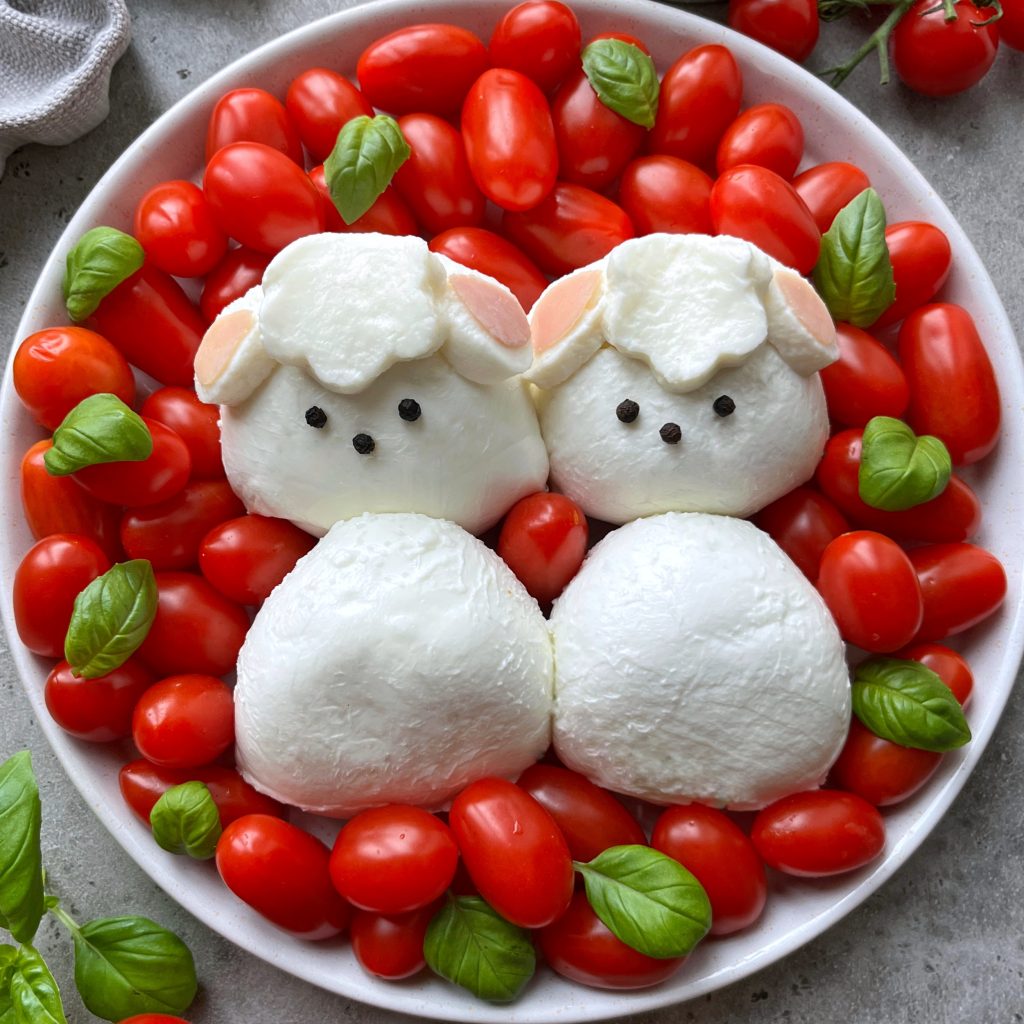 A plate with cherry tomatoes and basil leaves, featuring mozzarella cheese shaped like two sheep faces with black peppercorn eyes, surrounded by more mozzarella as their bodies.