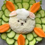 A cute Easter Cheese Ball salad shaped like a bunny, topped with white cheese and sliced ham, is surrounded by cucumber slices and carrot pieces arranged in a flower pattern on a white plate.